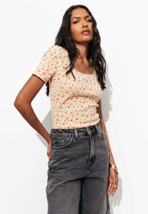 Femme aux longs cheveux foncés portant un haut court crème à manches courtes avec un petit imprimé floral rouge et un jean noir délavé taille haute, debout devant un fond blanc.