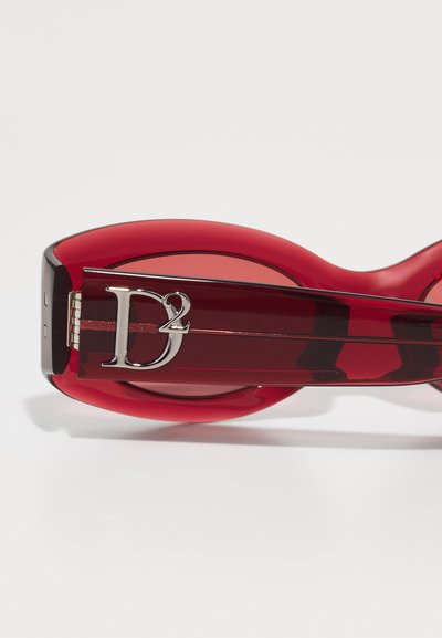 Red oval sunglasses featuring a glossy finish, a contrasting darker red arm, and a silver logo accent. Smooth texture with a bold design.