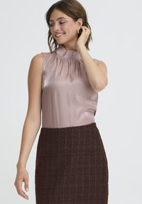 Blusa senza maniche di un leggero rosa pallido con un colletto alto arricciato, abbinata a una gonna scura in tessuto strutturato color marrone. Tessuto liscio con un delicato sheen.