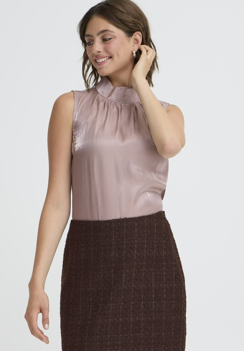 Blusa senza maniche di un leggero rosa pallido con un colletto alto arricciato, abbinata a una gonna scura in tessuto strutturato color marrone. Tessuto liscio con un delicato sheen.