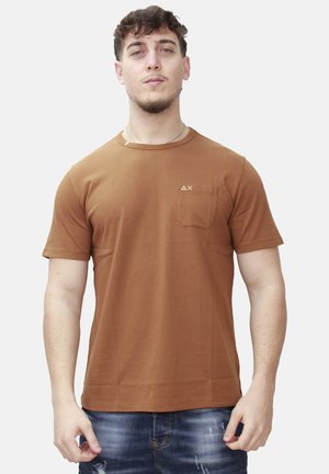 Uomo che indossa una t-shirt marrone a maniche corte con una tasca sul petto e un piccolo logo, abbinata a jeans blu strappati.