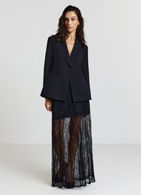 Schwarzer Blazer mit scharfen Revers über einem durchscheinenden schwarzen Spitzenkleid mit filigranen Mustern und fließender Silhouette. High Heels vervollständigen den Look.