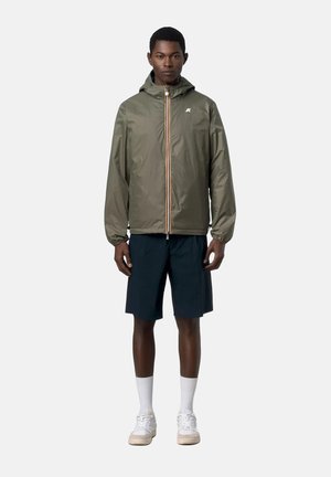 Uomo in piedi che indossa una giacca con cappuccio verde oliva, pantaloncini blu navy, calzini bianchi e scarpe da ginnastica bianche contro uno sfondo chiaro e semplice.