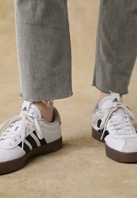 Weiße Sneakers mit braunen Gummisohlen und schwarzen Streifen, kombiniert mit ausgefransten grauen Jeanshosen in Knöchelhöhe.