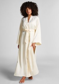 Vrouw met krullend haar, gekleed in een lange, crèmekleurige satijnen badjas die rond de taille is gebonden, staat blootsvoets tegen een effen lichte achtergrond.