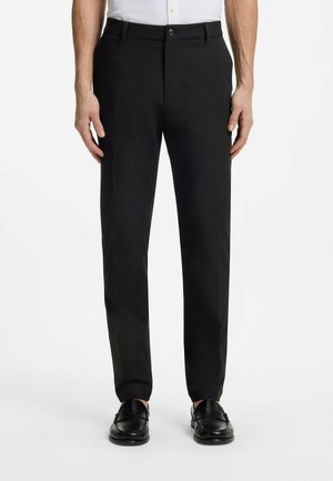 Mężczyzna noszący czarne, dopasowane eleganckie spodnie na guziki z szlufkami na pasek, w połączeniu z czarnymi mokasynami i białą koszulą.