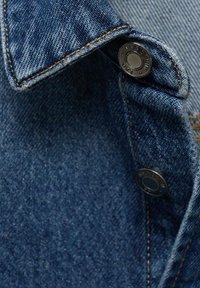 Denim tkanina v temno modri barvi z vidno šivanko. Ima ovratnik in pritisni gumb na vratu za zapiranje.