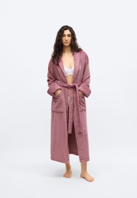 Γυναίκα που στέκεται ξυπόλητη, φορώντας ένα μακρύ ροζ μπουρνούζι πάνω από λευκά εσώρουχα, σε απλό ανοιχτόχρωμο φόντο.