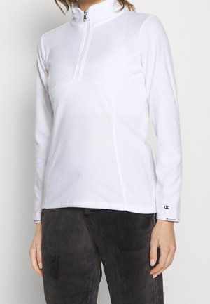 Frau trägt einen weißen langärmeligen Pullover mit halbem Reißverschluss und dunkle Hosen, mit sichtbaren Champion-Logos auf den Ärmeln.