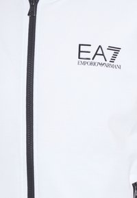 Giacca bianca con zip, caratterizzata da una cerniera nera e dal logo "EA7 Emporio Armani". Tessuto liscio con una superficie leggermente testurizzata.