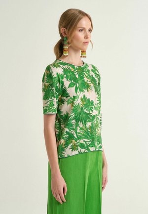 Donna con capelli castano chiaro raccolti in una coda di cavallo, indossa una maglia verde con stampa di foglie tropicali, una gonna verde vivace e orecchini a tassello colorati con perline.