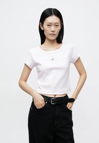 Light pink cropped t-shirt with ribbed texture and round neckline, featuring a small red logo on the chest. Worn with black denim pants and a belt.