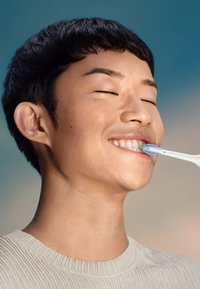 Spazzolino elettrico con manico blu e bianco. Persona che si lava i denti, con tono di pelle chiaro e capelli neri corti. Sfondo neutro.