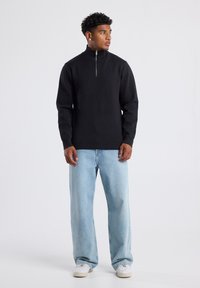 Black zip-up sweater with a high collar, paired with light blue relaxed-fit jeans and white sneakers. Smooth fabric, minimalist design.