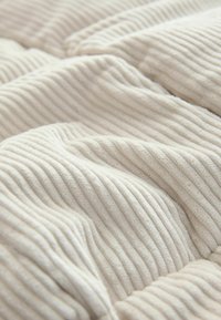 Weiches, geripptes cremefarbenes Gewebe mit einer plüschigen Textur, das subtile Falten und einen sanften Glanz aufweist und ein gemütliches Erscheinungsbild schafft.