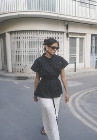 Crni top s pojasom i kratkim rukavima, uz dodatke na gumb, uparen s bijelom prozirnom suknjom koja sadrži uzorke listova i teksturirane akcentacije.