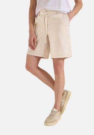 Person iført beige knælange shorts med snøring og beige sneakers, stående med krydsede ben og den ene hånd i lommen.