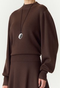 Person iført en brun langærmet sweater og nederdel med en sort-hvid vedhæng på en sort snor.