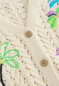 Gilet en tricot crème avec motifs torsadés, boutons en bois et fleurs brodées colorées sur un tissu texturé.