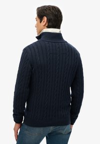 Navyblauwe gebreide trui met een hoge kraag, kabelpatroon, geribde manchetten en zoom. Materiaal lijkt zacht en textuur te hebben.