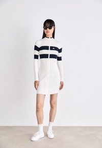 Abito bianco a coste con righe orizzontali blu scuro, colletto alto e zip frontale. Indossato con sneakers bianche e occhiali da sole neri.