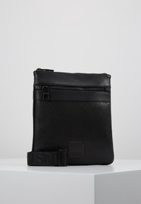 Černá kožená crossbody taška s herringbone vzorem, nastavitelným popruhem a zipovým uzávěrem. Obsahuje logo nášivku a hladkou texturu.