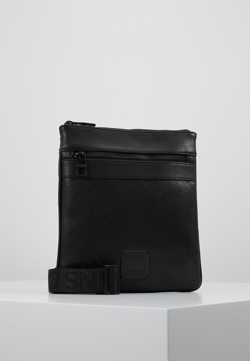 Černá kožená crossbody taška s herringbone vzorem, nastavitelným popruhem a zipovým uzávěrem. Obsahuje logo nášivku a hladkou texturu.