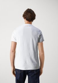 Camiseta blanca de algodón con mangas cortas, diseño ligeramente ajustado, cuello redondo y un detalle de logo elevado en la parte posterior, combinada con jeans de mezclilla oscura.