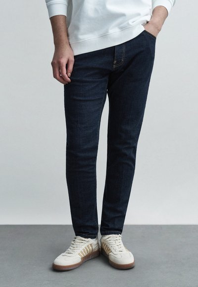 Donkerblauwe denim slim-fit jeans met tonale stiksels en zijzakken, gecombineerd met crèmekleurige sneakers met gouden accenten en textuurrijke details.