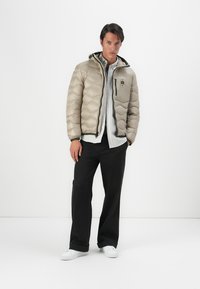 Puffer jacket in light taupe with quilted texture, front zipper, adjustable hood, and chest pocket. Paired with black trousers and white sneakers.