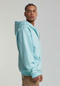 Lysblå hættejakke lavet af blødt stof, med lynlås, frontlommer og en rummelig hætte. Afslappet design med ribbede manchetter.