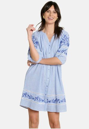 Mujer sonriente vistiendo un vestido suelto de color azul claro con bordados florales azules y mangas de tres cuartos, de pie con un brazo cruzado y la otra mano levantada.