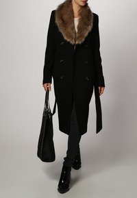 Kvinde iført en lang sort frakke med brun pelskrave, sorte bukser, sorte ankelstøvler og bærende på en stor sort studset håndtaske.