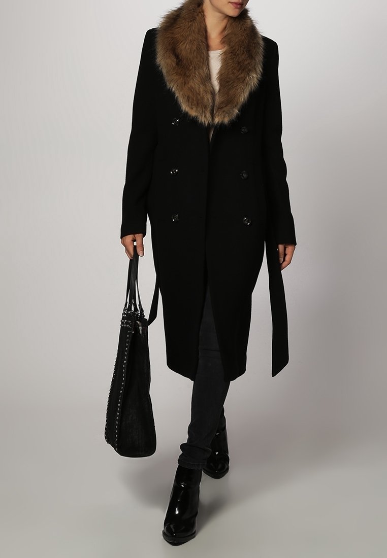 Kvinde iført en lang sort frakke med brun pelskrave, sorte bukser, sorte ankelstøvler og bærende på en stor sort studset håndtaske.