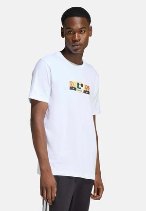 Giovane uomo che indossa una maglietta bianca Adidas con una grafica di una palla sportiva e pantaloni neri con strisce bianche, in piedi davanti a uno sfondo semplice.