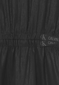 Černé šaty s texturovaným, pomačkaným designem, které mají na pase řasení a jsou opatřeny štítky Calvin Klein připevněnými k látce.