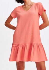 Vestido coral rosa con mangas cortas, escote en V y una tela texturizada. Presenta un dobladillo acampanado en capas para un ajuste relajado.