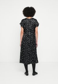 Schwarzes Kleid mit weißen Punkten, kurzen Ärmeln, mittlerer Länge und glatter Textur. Die Rückseite zeigt einen einfachen Ausschnitt und eine fließende Silhouette.