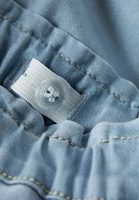 Lyseblå denimstoff med rynket tekstur. Har en hvit vevd etikett med en gjennomsiktig knapp og kontrasterende beige søm.