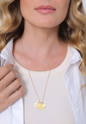 Goldkette mit einem strukturierten runden Anhänger und zwei Muschelakzenten, getragen über einem hellen gerippten Oberteil unter einem weißen Hemd.