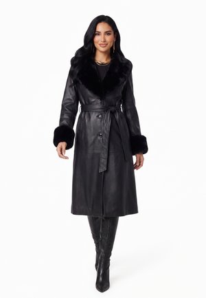 Donna che indossa un lungo cappotto di pelle nero con colletto e polsini in pelliccia, vita cinturata, abbinato a stivali neri fino al ginocchio.