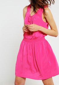 Ärmlös rosa klänning i slät tyg, med en rynkad elastisk midja och rosett-detalj vid halslinningen.