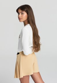 Camicia bianca a maniche lunghe con dettagli stampati, abbinata a pantaloni corti beige a pieghe. Il modello ha lunghi capelli castani e sta guardando di lato.