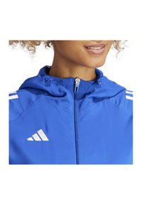 Blauwe ritsjack met capuchon, lichtgewicht stof, witte accentstrepen en een logodetail op de linkerborst.