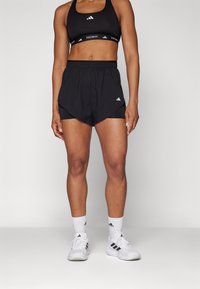 Sorte sportshorts med lagdelt design over tætsiddende indershorts, accentueret med hvid branding. Parret med hvide sneakers og sokker.