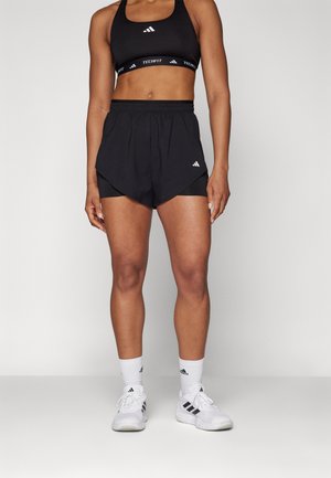 Schwarze Sportshorts mit geschichtetem Design über eng anliegenden Innenshorts, akzentuiert mit weißer Markenkennzeichnung. Kombiniert mit weißen Sneakern und Socken.