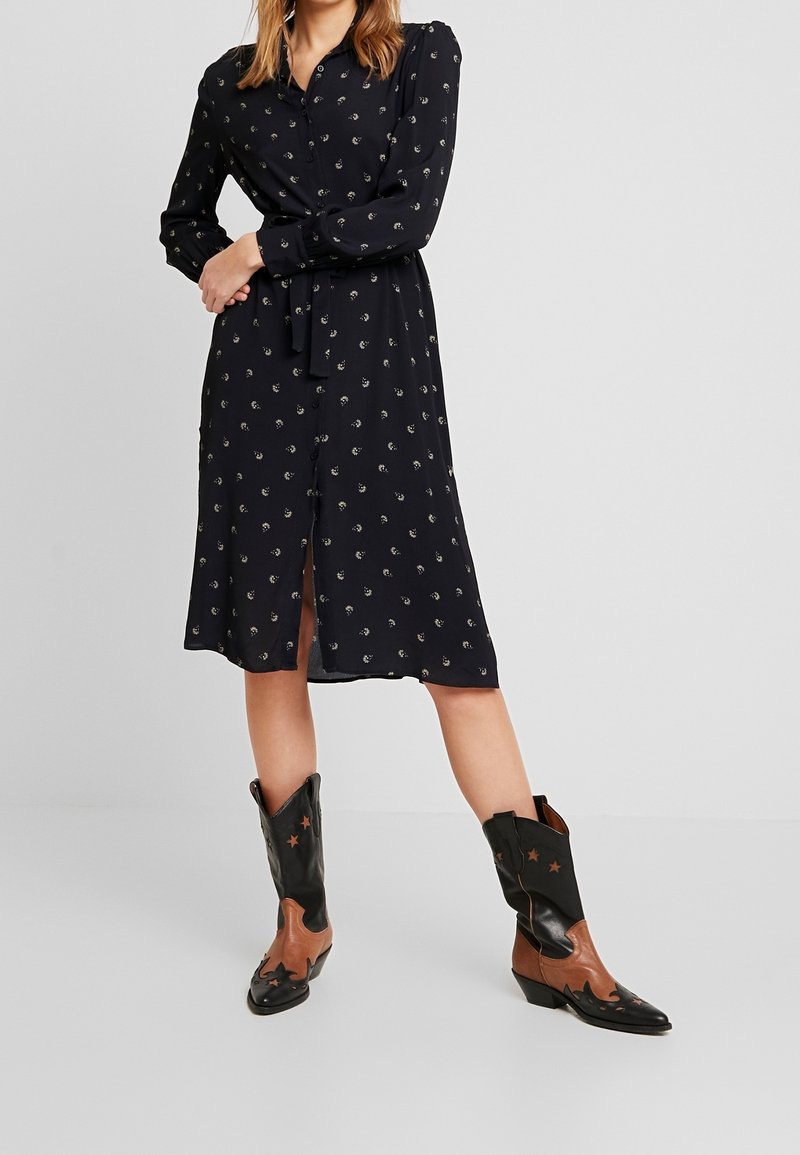 Mörkblå långärmad klänning med små guldmönster, framknappar och knytmidja, matchad med svarta och bruna cowbojboots med stjärndetaljer.