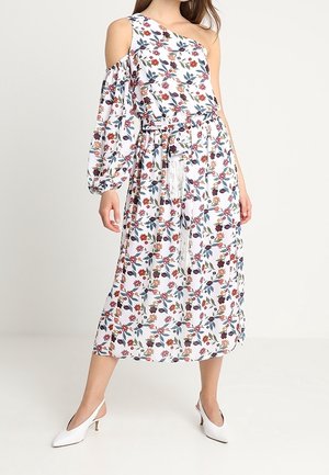 Kobieta w białej, kwiecistej sukience midi na jedno ramię z bufkami na rękawach i wiązaniem w talii, w białych butach na obcasie z paskiem z tyłu, na neutralnym tle.