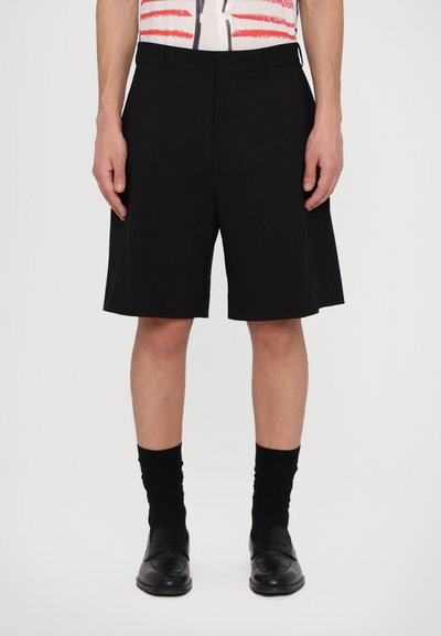 Shorts noirs ajustés en tissu lisse, avec une coupe droite, une taille et des poches avant. Portés avec des chaussettes noires et des chaussures.