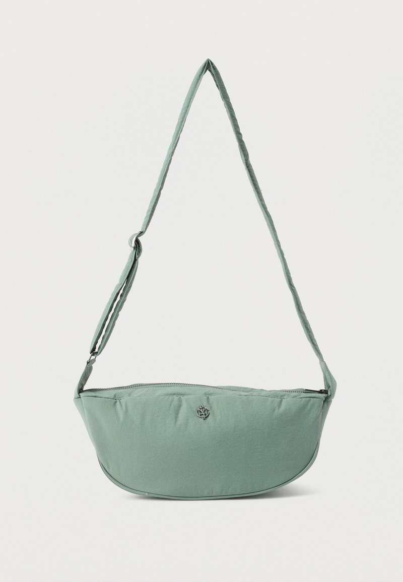 Ανοιχτό πράσινο ύφασμα τσάντα χιαστί με ρυθμιζόμενο ιμάντα και μικρό μεταλλικό λογότυπο στο μπροστινό μέρος, παρουσιάζεται σε λευκό απλό φόντο.